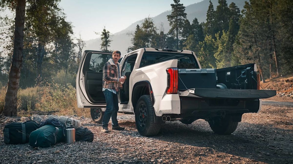 A man unloads camping gear in the forest from a Toyota Tundra with its tailgate down and rear door open.