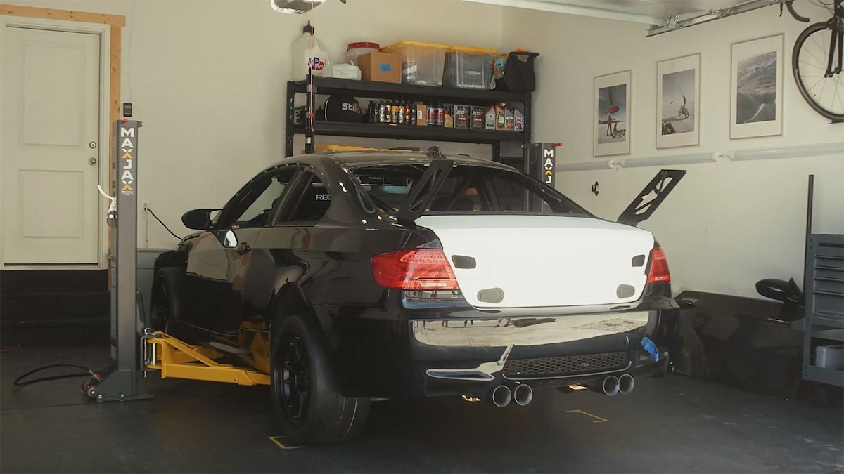 A black BMW M3 sits on a lift in a small garage. The trunk is white.