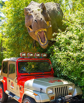 A red and silver Jeep with the Jurassic Park logo on the door parked next to trees with a dinosaur emerging from the leaves