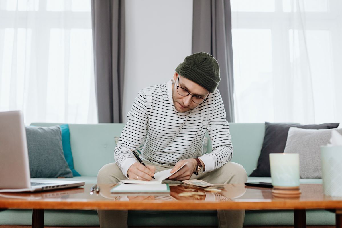 A man writes his budget in a notebook, while sitting on a couch. 