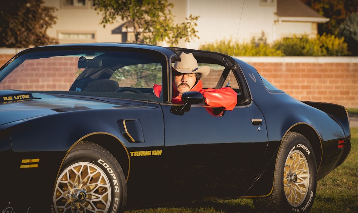 Johnny, wearing a tan cowboy hat and red leather jacket, looks just like Burt Reynolds sitting in the driver's seat of his black Pontiac Trans Am