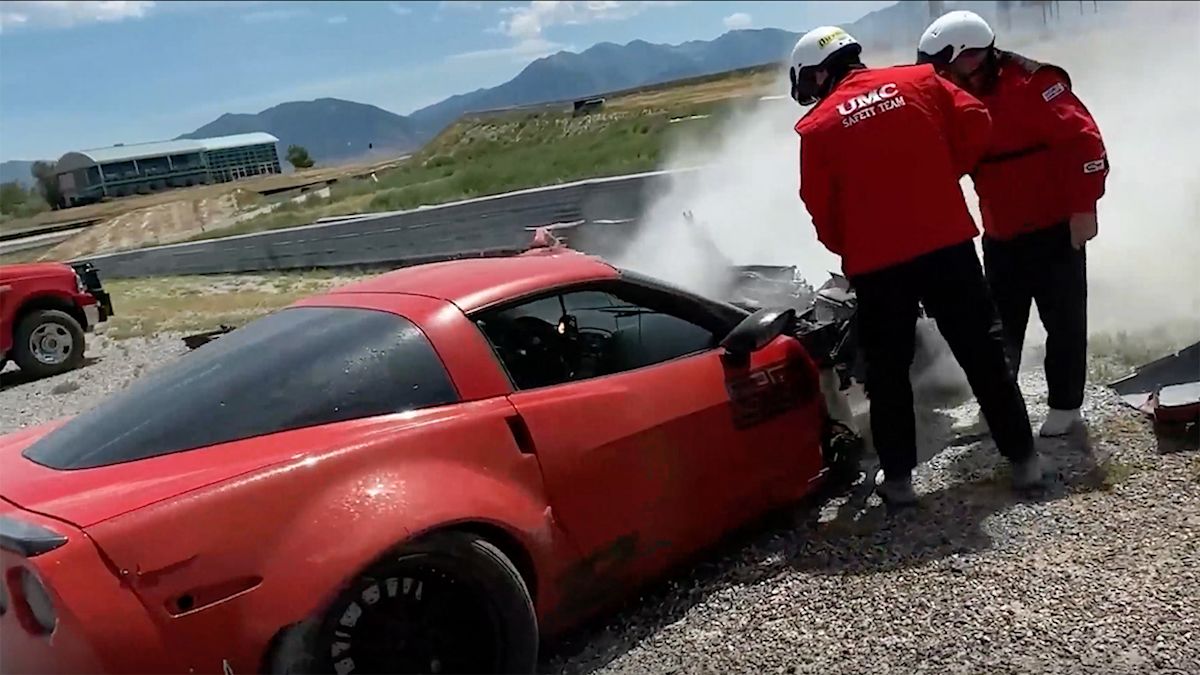 A red Corvette on a racetrack with its front end smashed in and smoking. Two first responders attend to it.