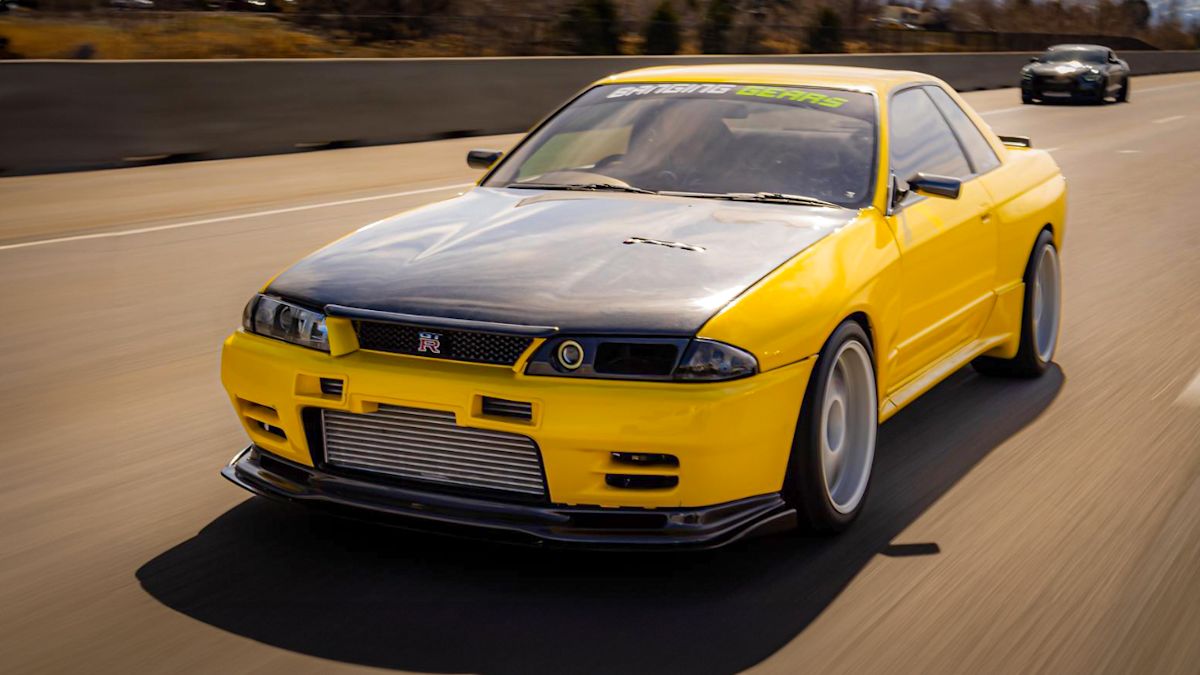 A yellow Nissan Skyline R32 with a black hood speeds around a closed race track, with another supercar close behind