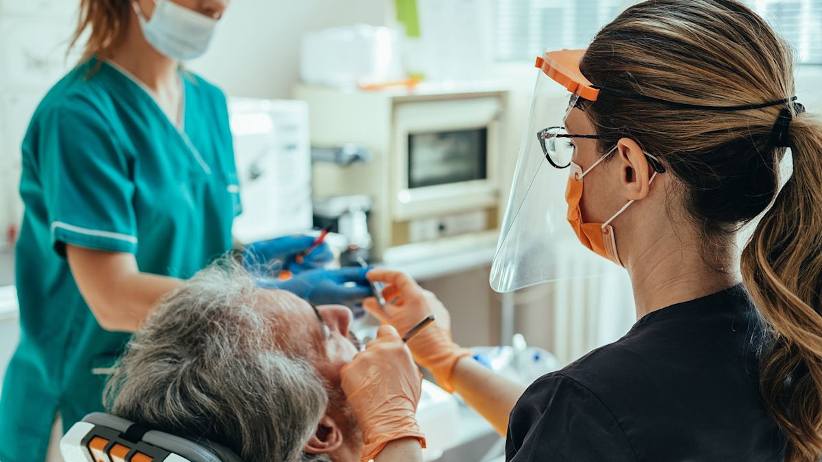 A dental assistant hands a dentist a tool as she works on a gray-haired male patient reclined in a dental chair.
