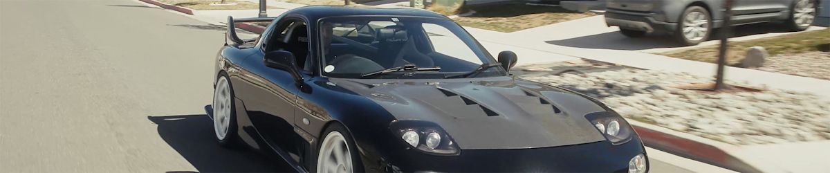 A black Mazda RX7 drives down a road lined with townhomes.